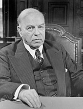 An older white man in a pinstriped three piece suit sits facing the camera, his right hand resting in front of him on a table.