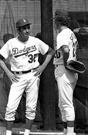 "Two men wearing Los Angeles Dodgers uniforms and caps; one is older with hands on his hips and listens to the younger man make conversation."