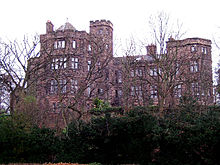 Seen from a low angle through the branches of trees is a substantial stone house with two-three storeys, bay windows and some castellation.