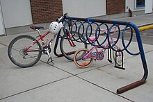 An open A-frame of thick steel tubing. Attached underneath the crossbar is a spiral of thinner metal tubing. It is painted blue, but much of the paint has flaked off, leaving rust. A mountain bike and a child's bike are parked in it and two abandoned D-locks hang from it. In the background is a brick-and-plaster building and behind the rack, to the right, is a car parking space.