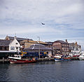 Passenger train passing between fishing boats and buildings in 1994, with Concorde overhead