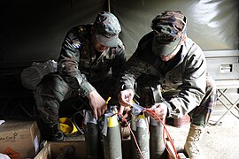 A group of 105 mm artillery shells with plastic explosive stuffed into their fuze pockets to act as booster charges. Each of the five shells has been linked together with red detcord to make them detonate simultaneously. To turn this assembly into a booby trap, the final step would be to connect an M142 firing device to the detcord and hide everything under some form of cover e.g. newspapers or a bed-sheet.