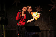 Photograph of the siblings almost back-to-back in front of a microphone stand, with Laser holding a cat-shaped electronic piano.