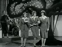 A black and white image of three women with arms interlocked standing in front of a wooden tree-shaped prop. Men with trombones and saxophones are visible in the background.