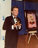 A smiling, formally clad man holding a Grammy Award.