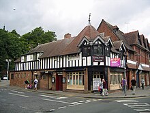 A two-storey building seen from the corner; the lower storey is brick and the upper storeys timber-framed. The left side extends further than the right and contains the public conveniences, with two doorways on the ground floor and a long dormer above. On the corner is a doorway with windows on each side; above are three windows extending round the corner, each with a gable. Behind these is a tiled spire.