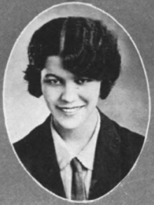 A smiling young Latina woman with dark wavy hair cut short; wearing a menswear-style suit and necktie