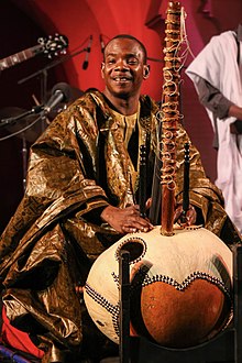 Photograph of Diabaté, seated onstage in a grand boubou robe, playing kora