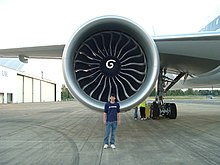 Aircraft engine, forward-facing view with a Boeing engineer in front to demonstrate the engine's size. The engine's large circular intake contains a central hub with a swirl mark, surrounded by multiple curved fan blades.