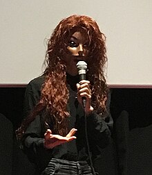 Narcissister wearing a mask and gesturing as she speaks. She is standing in front of a projection screen.