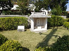 Photograph of a small white concrete shrine opened on the front, in a small grass covered space, surrounded with low shrubs. There is an explanation panel on the left of the shrine and an incense burner and a stele saying "Hi-nu-kan" inside the shrine.