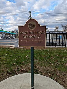 Sign in a park that says "Anne Sullivan Memorial, Feeding Hills, Massachusetts, Funded by the Community Preservation Act Committee" under the Town of Agawam seal.