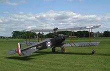 Left rear quarter view of restored dark brown Avro 504 biplane in Royal Air Force markings, on grass with trees on the horizon, and a partly cloudy sky.