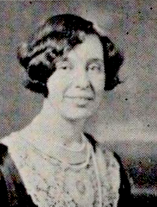 A smiling white woman with wavy dark hair, wearing a dark dress with a light lace yoke detail, and pearls