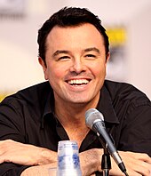 A man with black hair, and tan skin with a black shirt on, leans forward while laughing into a microphone.