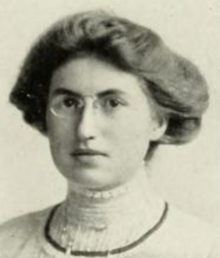 A young white woman with dark hair in a bouffant updo, wearing eyeglasses and a high-collared white blouse with lace trim; also a strand of dark beads