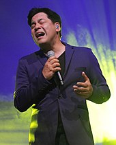 An image of Nievera wearing a navy blue suit singing into a hand-held microphone