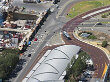 View from a skyscraper of a curved roof that fully covers the station. Next to the station is a busway that enters and exits Elizabeth Quay bus station. East of the station are buildings being constructed at Elizabeth Quay.