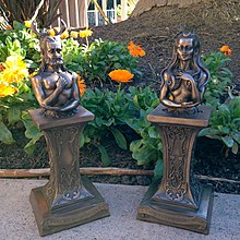 Two brownish metal bust statues on pedestals. On the left, a bearded, aged man with deer horns representing the Horned God. On the right, an aged woman with a waning crescent above her forehead representing the senescent "crone" form of the Triple Goddess.
