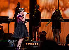 Kelly Clarkson performing in a dark purple dress against an orange-lit LED screen background