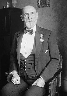 A bearded man, seventy years old, sitting, dressed in three-piece suit with bow-tie and silver medal hanging from left lapel.