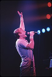 A short-haired man singing into a microphone while holding his left hand high in the air