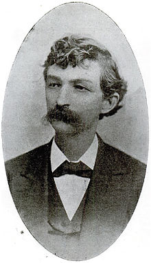 A man with curly, dark hair and a thick, dark mustache wearing a black jacket and tie and white shirt