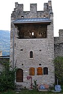 Tower in the centre of a stone wall. At the bottom left of the tower there is a door and then, on its right, two small wooden shutters. In the middle of the tower wall there is a small window and at the top of the wall a balcony is protected by a roof. The top of the tower is crenellated.