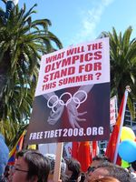 A crowd of people protesting against the 2008 Olympics. In the foreground there is someone holding up a white poster which says "What Will The Olympics Stand For This Summer?" in red capital letters at the top and at the bottom it shows an image of a pair of hands held together by the Olympic rings as if they were handcuffs.