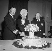 A man in naval uniform and a woman wearing a hat cut into a cake labeled Operation Crossroads, and shaped like a mushroom cloud, while another naval officer looks on.