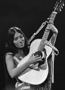 Black and white photo of Buffy sainte-Marie from the waist up playing an acoustic guitar