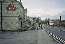 Gray stone building on the left with a pub sign outside it. A road is central to the picture with a white coloured building on the right.