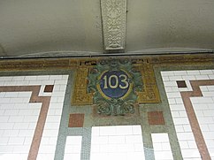 A cartouche on the wall of the 103rd Street station. There are white ceramic tiles, separated by bands of green mosaic tiles, as well as scattered sections of salmon mosaic tiles. At top center is a carved cartouche, surrounding a blue oval with the number "103" in white text.