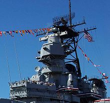 A large tower on a ship, on top is a radar antenna with an enclosed lookout space below the radar and a ship bridge below the enclosed space. Several people can be seen standing on the top of the bridge.