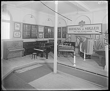 A photograph of a piano exhibition by Hornung & Møller. The picture features the corner of a rectangular room. Two walls are visible. Three upright pianos stand against the left wall which features glass cases containing piano actions, as well as what appears to be framed diplomas. A window is located to the very left with daylight visible through the curtains. The right wall features another upright piano, grand piano parts, more encased piano actions, framed photographs of pianos, as well as a large sign displaying the words “Hornung & Møller A/S, Kgl. hof-pianofabrik, grundlagt 1827.”. On the floor in front of the corner is another upright piano, as well as two grand pianos. To the very right is a display case and a plant on top of a pedestal. The center of the room features a lowered floor and in the very center is a coloumn which appears to support beams in the ceiling above. The ceiling also features multiple electric luminaries.