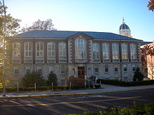 A tall brick building with a hipped roof, central pavilion and stone foundation seen from across a road with the sun just beyond the left of the image.