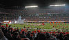 A large stadium that appears to be nearly full with a celebration taking place on the field with fireworks, athletics and others carrying flags.