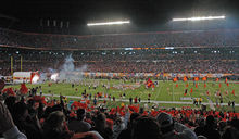 Distant figures rush onto a darkened football field carrying orange flags. In the foreground are the heads of spectators.