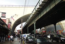 A view of the sidewalk on 31st Street, looking north toward the station house of the Astoria–Ditmars Boulevard BMT station, at 31st Street and 23rd Avenue in Astoria, Queens. The Hell Gate Bridge's concrete approach viaduct rises above the station in the background.