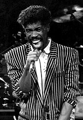 A dark-skinned man in a pinstripe jacket singing into a microphone