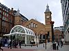 The end of a brick building with arched windows and sloping roofs lies between two towers with steeples. In front of this is a white metal and glass structure. People are standing and walking in the street in front.