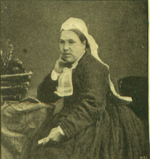B&W portrait photo of a seated middle-aged woman in a dark dress, wearing a white head scarf and a white lace kerchief.