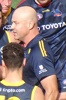 A fair-skinned bald man in a navy polo speaking to football players