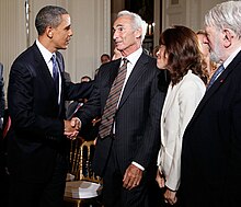 A younger African-American man shakes hands with an older Jewish-American man; both of them are wearing dark suits. Next to the older man is a woman wearing a white suit, facing away from the camera