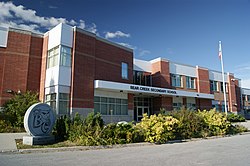 Main entrance of the school. A circular rock is shown with the school logo next to a bush wall. The Canadian flag is seen.