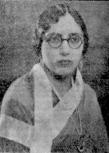 A South Asian woman with hair parted center and dressed in marcelled waves, wearing round glasses and a silk shawl with a pendant necklace and earrings