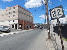 A sign with the number "22" in black on a white shield, itself on a black background, on a slightly skewed metal post stands at front right. Across a road are some low multistory urban buildings