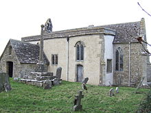 A small stone church seen from the south-east with a porch to the left, a protruding rendered south aisle in the middle, and a short chancel to the right. On the west gable is a bellcote, and in front of the church is a churchyard cross