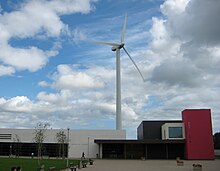 Picture of a building taken during the day - it is white on the left, and black on the right, with one narrow segment that is coloured red. There is a windmill in the background. The picture is taken from afar. The sky is blue with white clouds.