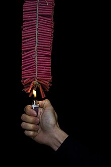 A string of red firecrackers. A young man holds a flame to set it off.